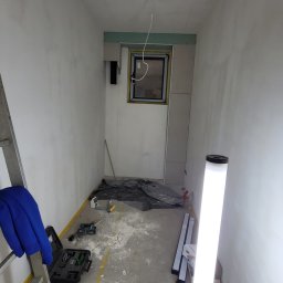 Usługi leśne - Wąskie pomieszczenie w trakcie remontu: ściany po szpachlowaniu, okno z żółtą ramą, drabina z niebieską tkaniną, lampa budowlana i narzędzia na podłodze.