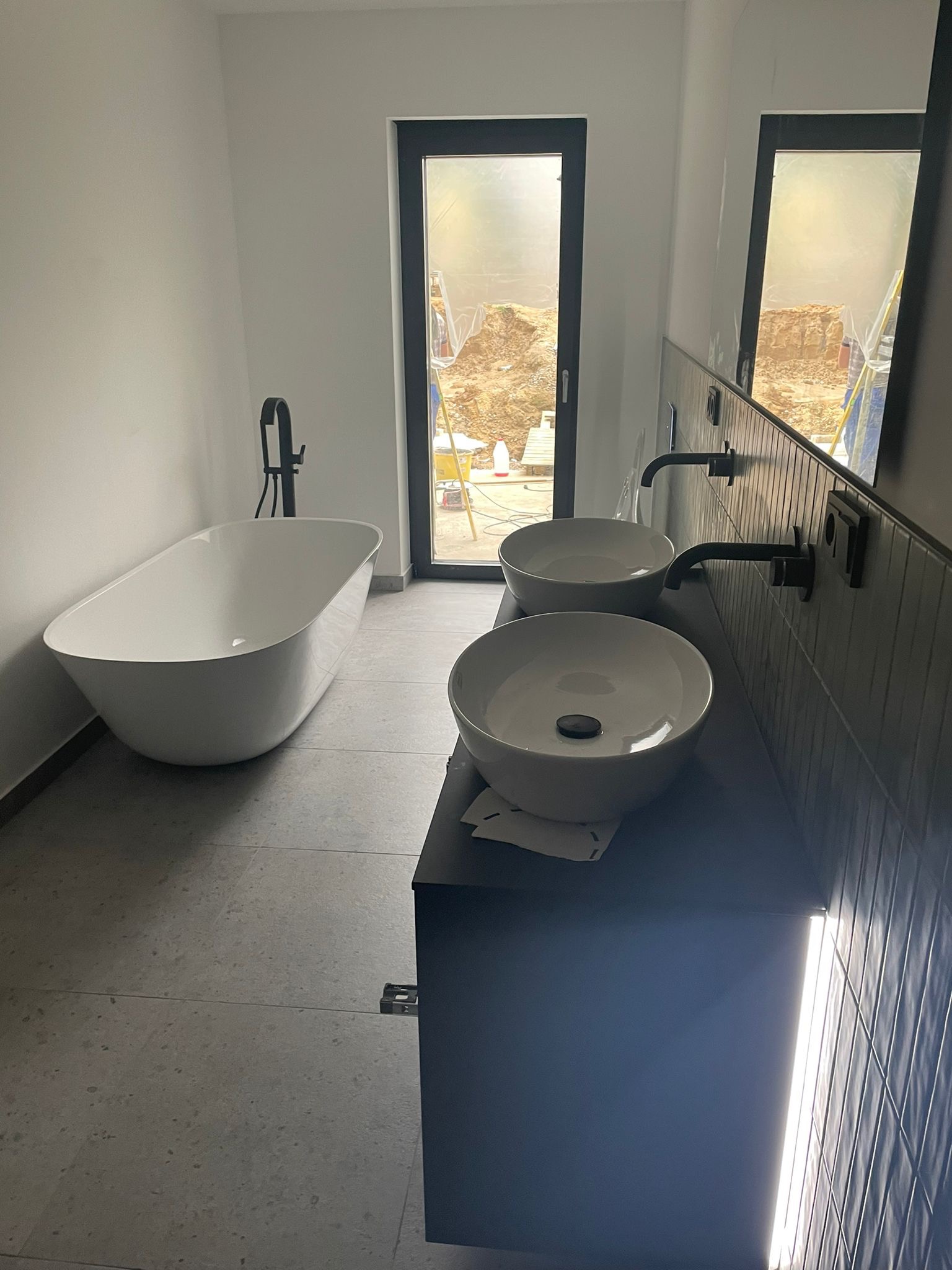 Nowoczesna łazienka w trakcie wykańczania, z wolnostojącą białą wanną, podwójną umywalką nablatową na czarnym blacie i czarną armaturą; widok przez okno na plac budowy z hałdą ziemi.