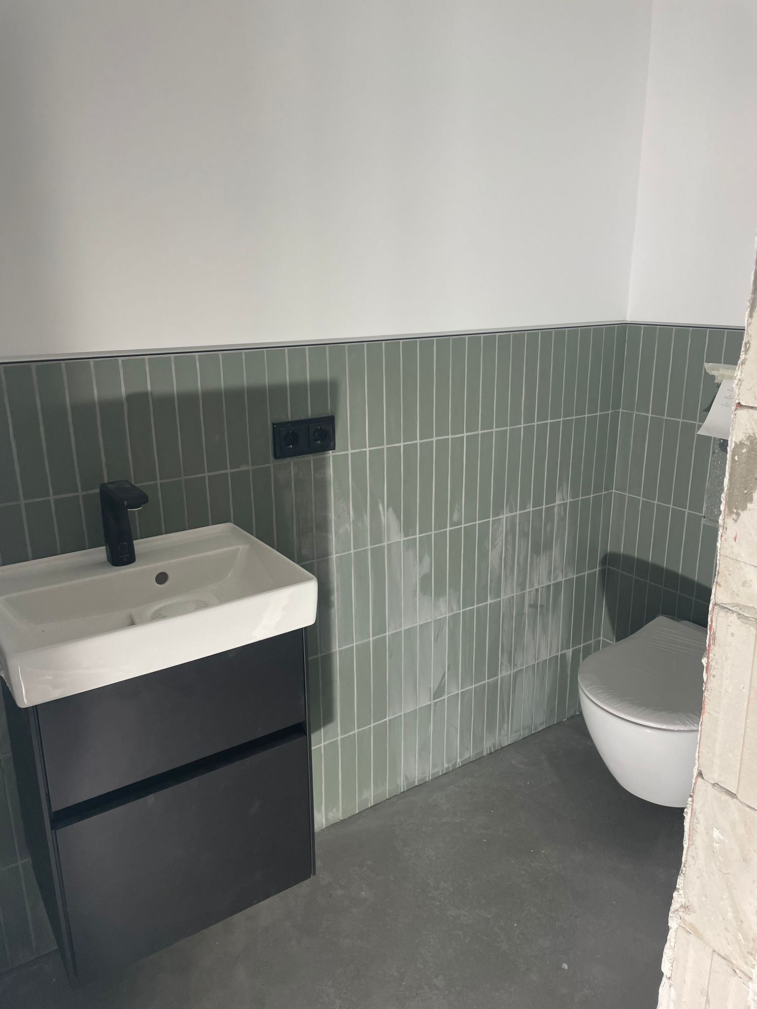 Nowoczesna łazienka w trakcie wykończenia: pionowe, zielone płytki na ścianie, czarna armatura przy białej umywalce z szafką, widoczny fragment toalety i surowy stan wykończenia.