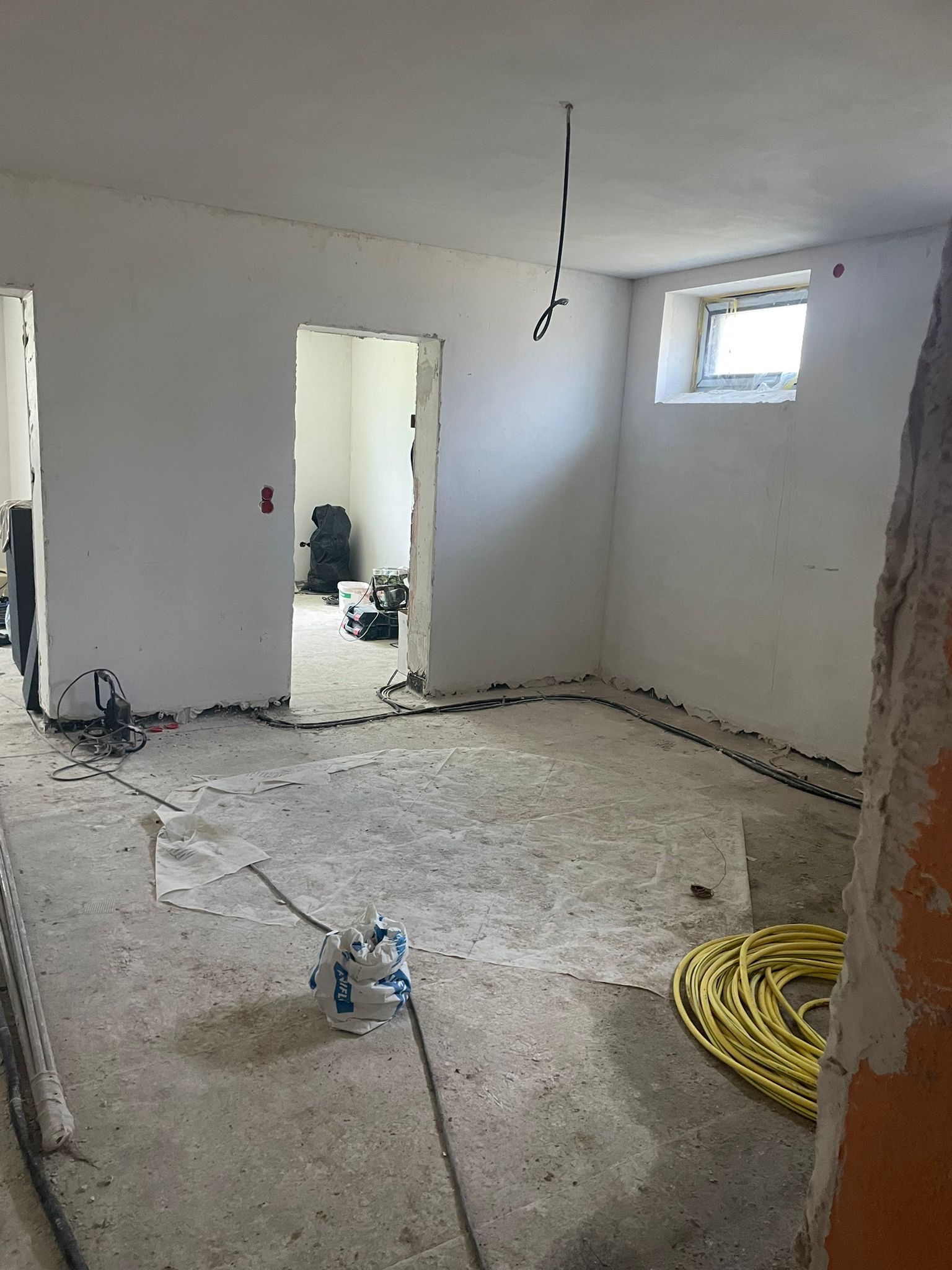 Surowe wnętrze w trakcie remontu: białe ściany, betonowa podłoga zabezpieczona folią, widoczne kable elektryczne, worki z materiałami budowlanymi i zwój żółtego kabla. Widok z perspektywy wejścia...