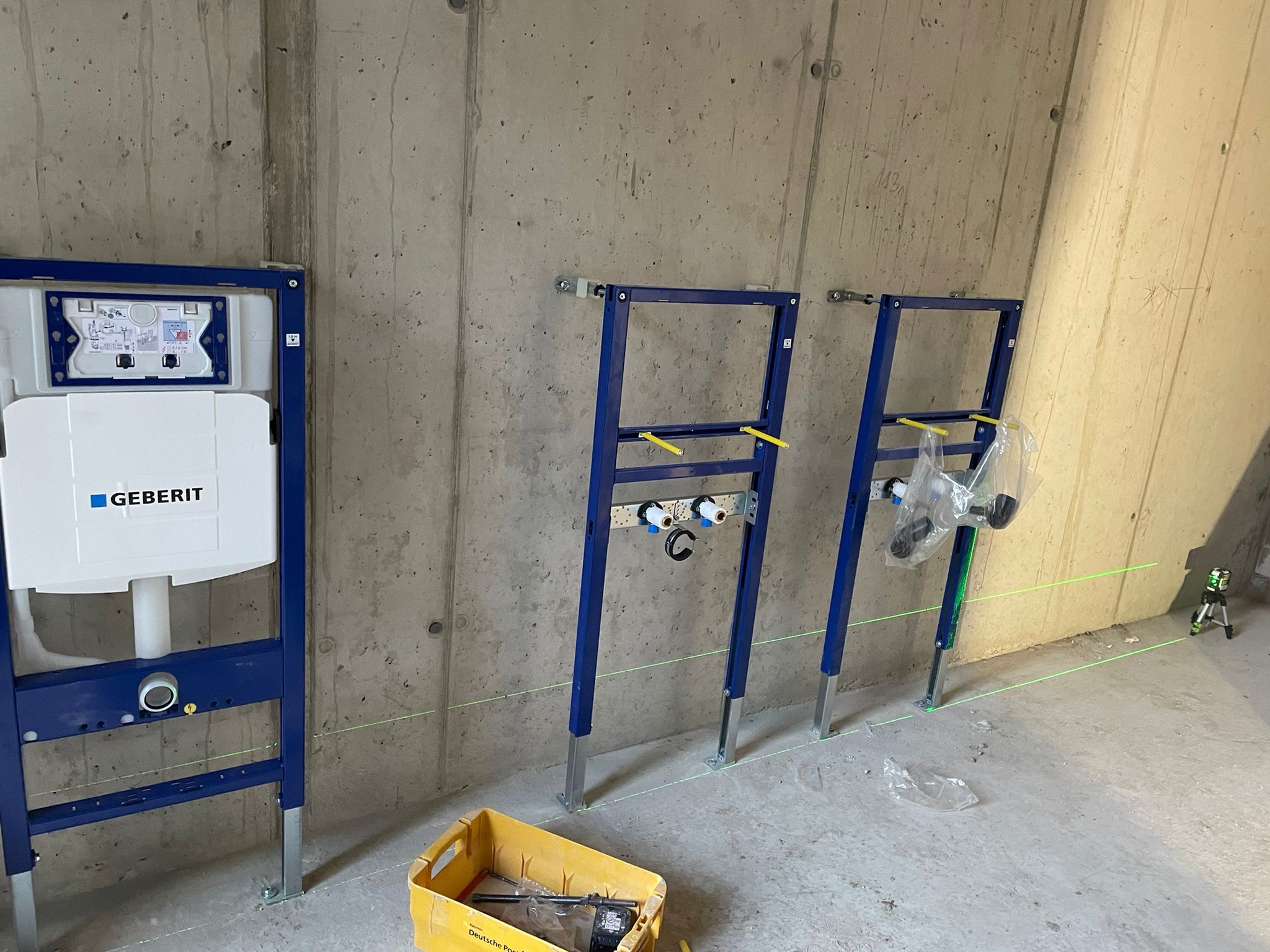 Stelaże podtynkowe Geberit zamontowane na surowej ścianie z betonu, widoczny laser krzyżowy wyznaczający poziom, żółty pojemnik z narzędziami na podłodze.