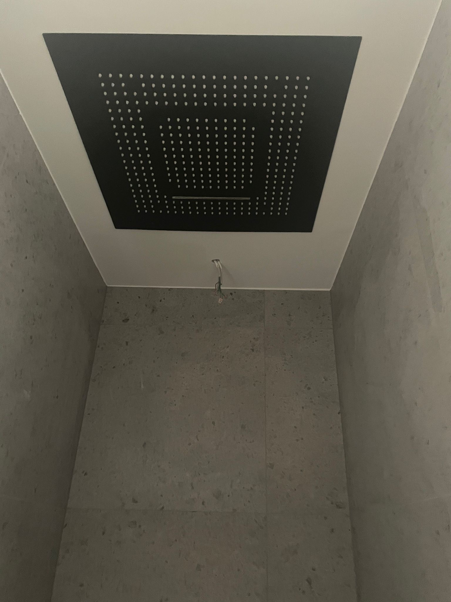 Nowoczesna kabina prysznicowa z kwadratową, czarną deszczownicą sufitową i szarymi płytkami imitującymi kamień, widoczne wystające przewody.
