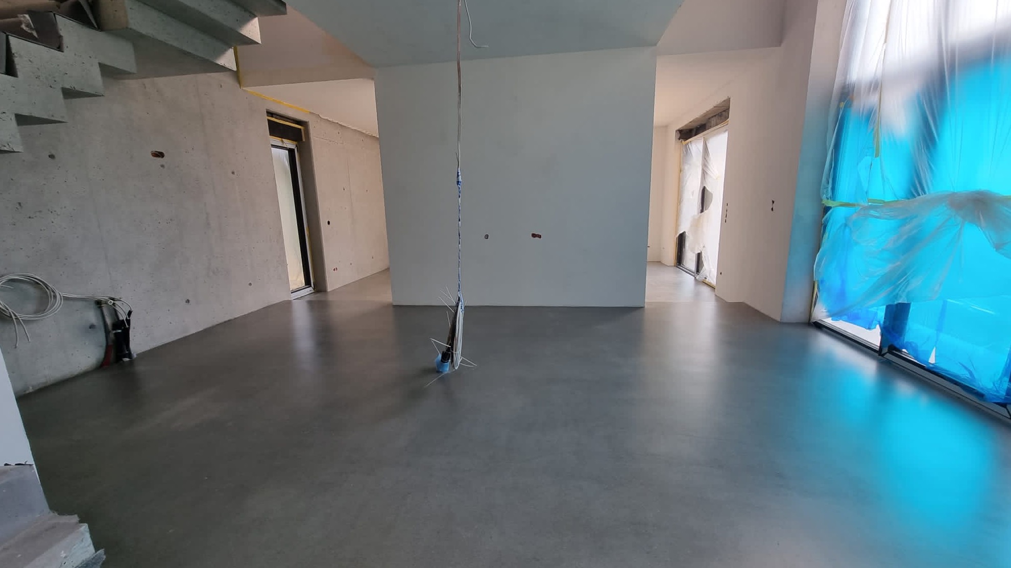 Surowe wnętrze domu w trakcie remontu: betonowe ściany, wylewka na podłodze, okna zabezpieczone folią malarską, widoczne przewody elektryczne. Schody betonowe w lewym górnym rogu.