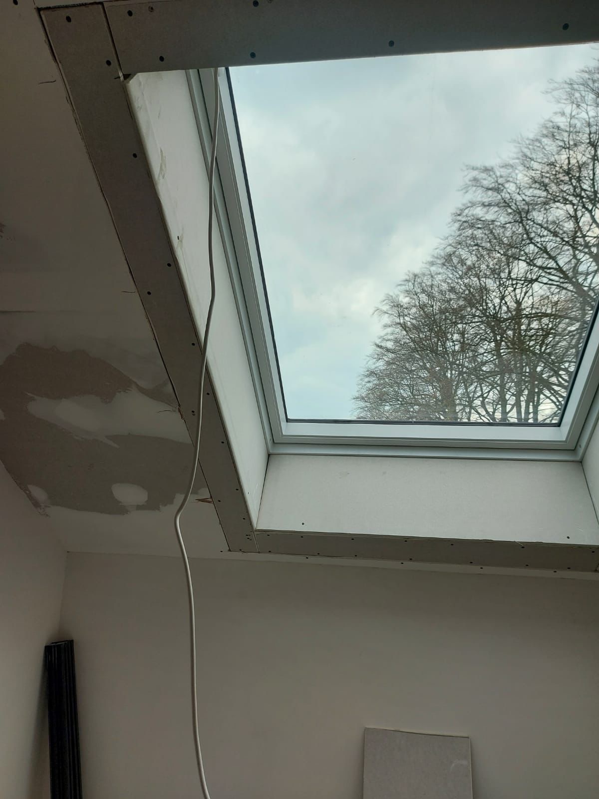 Wnętrze w trakcie wykańczania z zamontowanym oknem dachowym, widoczne płyty gipsowo-kartonowe na suficie i ścianach, częściowo wyszpachlowane, z widokiem na drzewa przez okno.