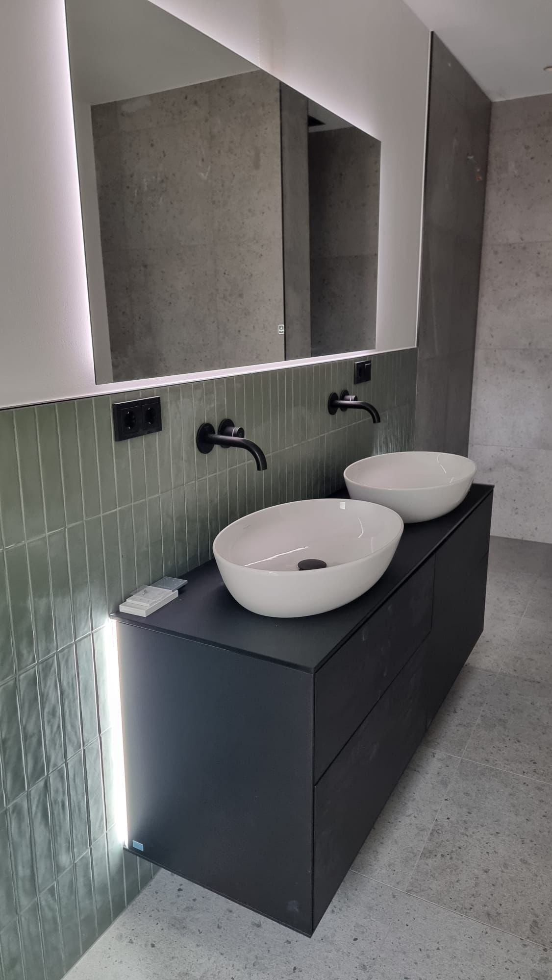 Nowoczesna łazienka z dwoma białymi umywalkami nablatowymi na czarnej szafce, podświetlane lustro z ledowym oświetleniem, zielone pionowe płytki na ścianie i czarne baterie.