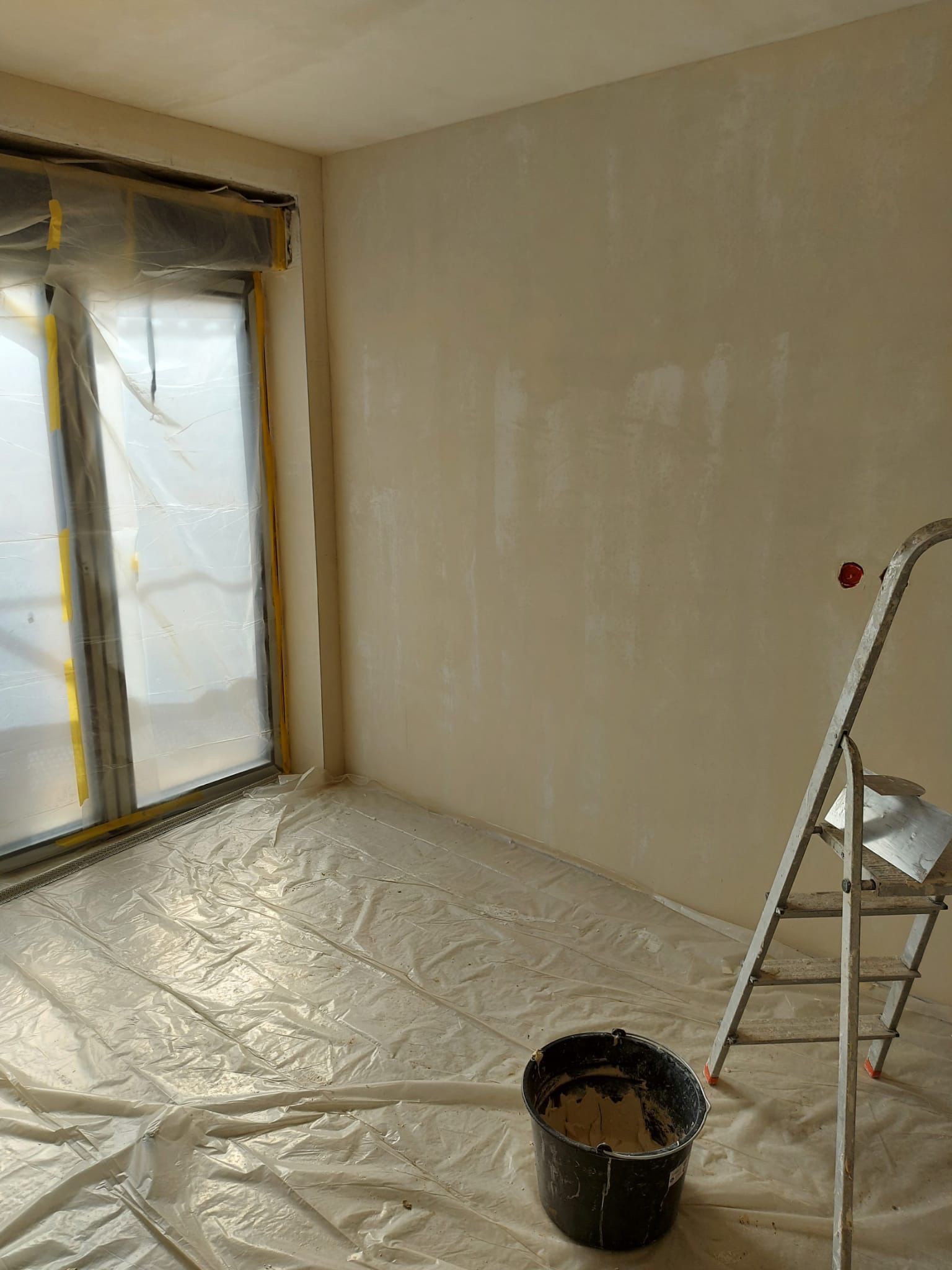 Pokój w trakcie remontu: ściany przygotowane do malowania, podłoga i okno zabezpieczone folią, drabina i wiadro z zaprawą w pomieszczeniu.