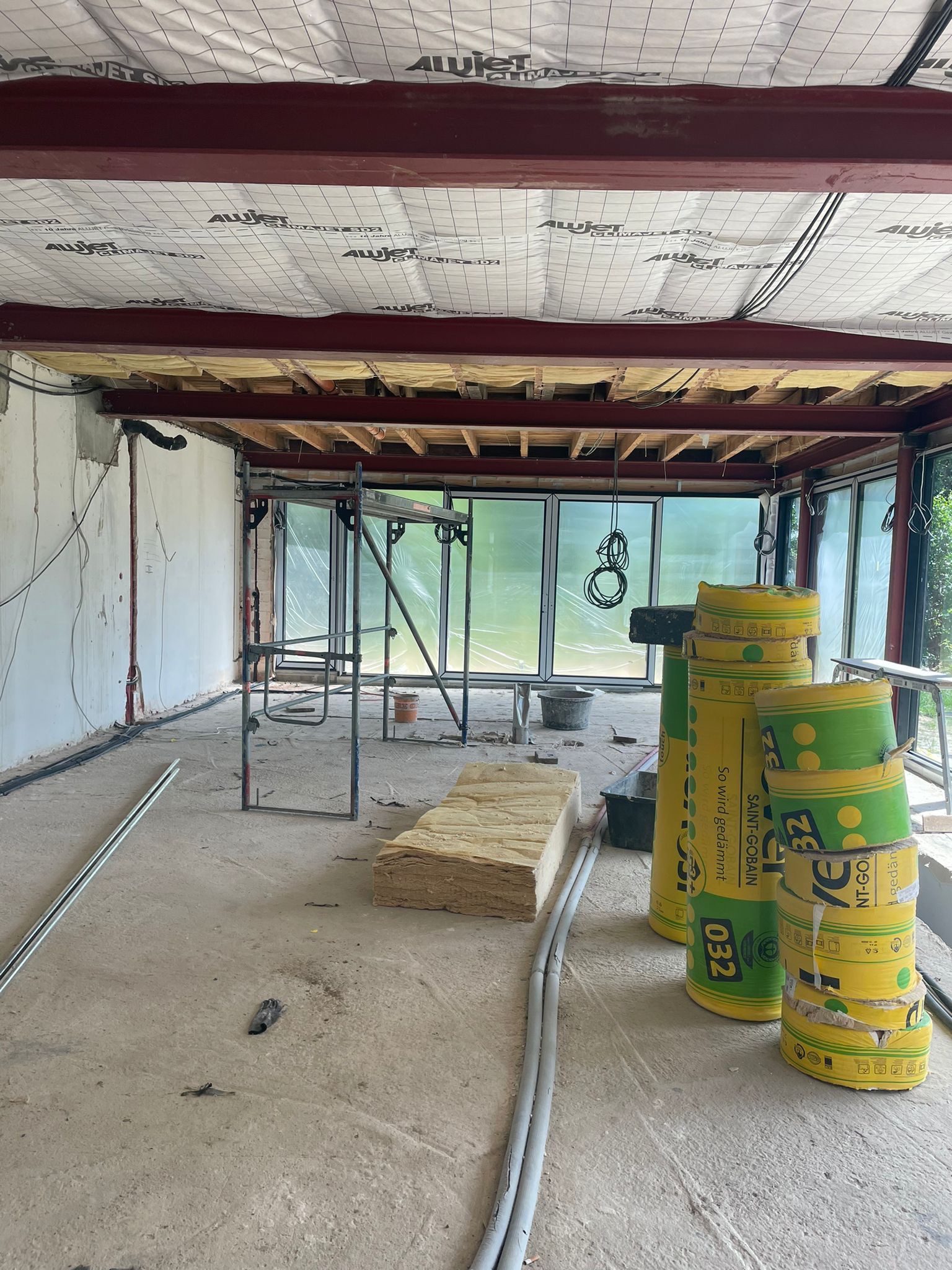 Wnętrze budynku w trakcie ocieplania: widoczne stalowe belki stropowe z izolacją, rusztowanie, rolki wełny mineralnej w folii, przewody elektryczne i surowe ściany.