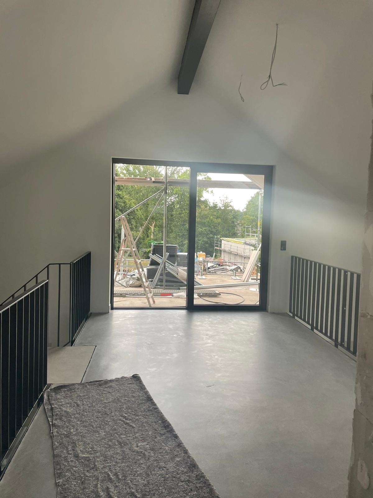 Wnętrze domu w stanie deweloperskim z widocznym betonowym podkładem podłogowym, czarnymi balustradami schodowymi i dużym przesuwnym oknem z widokiem na zewnątrz, gdzie widać rusztowania i materiały...