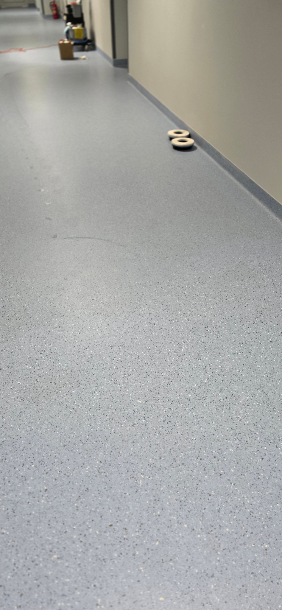 Długi korytarz z szarą podłogą w plamki, w tle maszyna czyszcząca podłogi i ślady na podłodze.