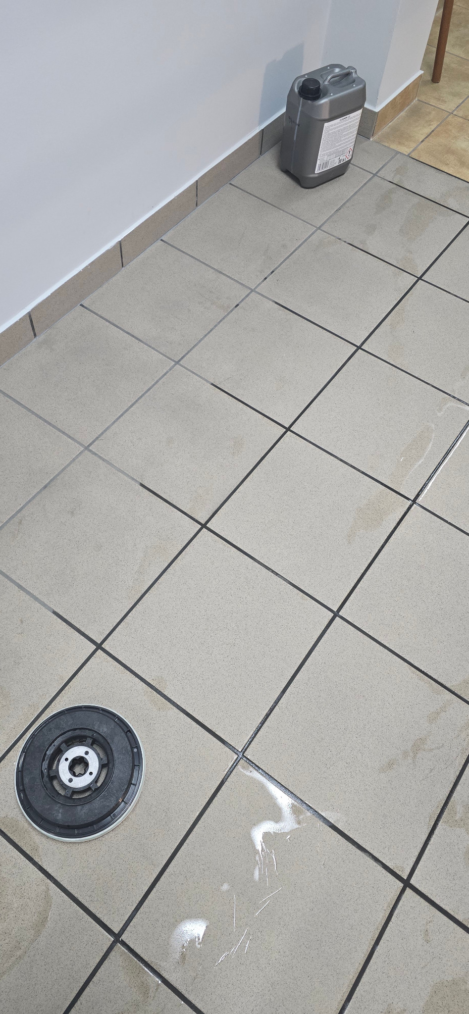 Maszyna do czyszczenia podłóg na kafelkach, obok kanister z płynem czyszczącym i resztki płynu na podłodze.