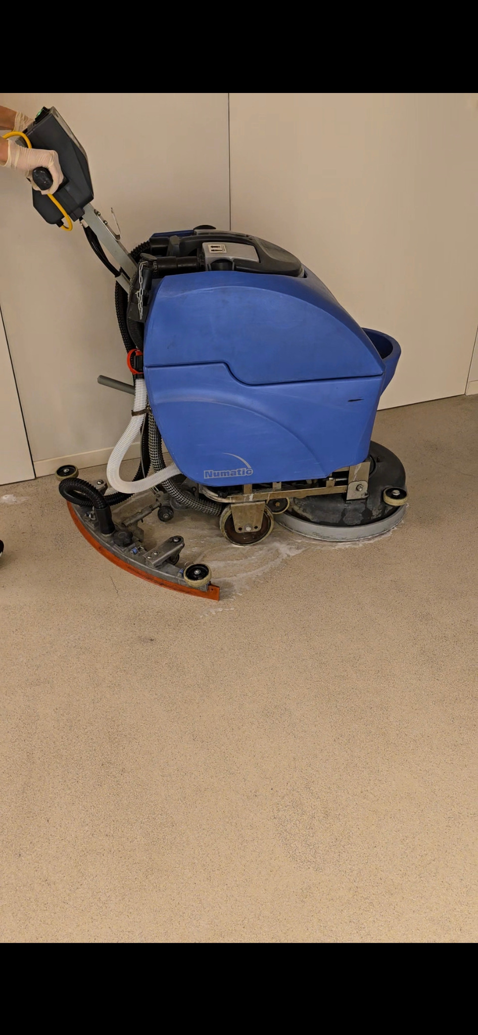 Automat szorujący Numatic w trakcie pracy na beżowej podłodze, widoczna piana czyszcząca i osoba w rękawicach ochronnych obsługująca maszynę.