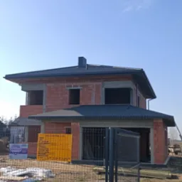 Niedokończony budynek dwukondygnacyjny z ciemnym dachem i widoczną konstrukcją z cegieł, otoczony ogrodzeniem z siatki i tablicą informacyjną budowy.