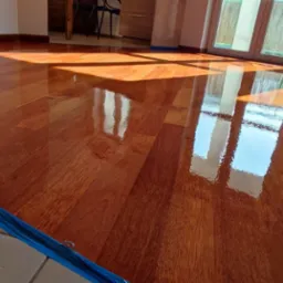 Błyszcząca, świeżo polakierowana podłoga z drewna egzotycznego, zabezpieczona niebieską folią malarską przy krawędziach, z wyraźnymi refleksami światła słonecznego padającego przez okno.