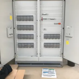 Otwarta szafa rozdzielcza z zamontowanymi bezpiecznikami i elementami sterującymi, widoczne kartony i elementy montażowe na podłodze, w tle instalacje pod sufitem.