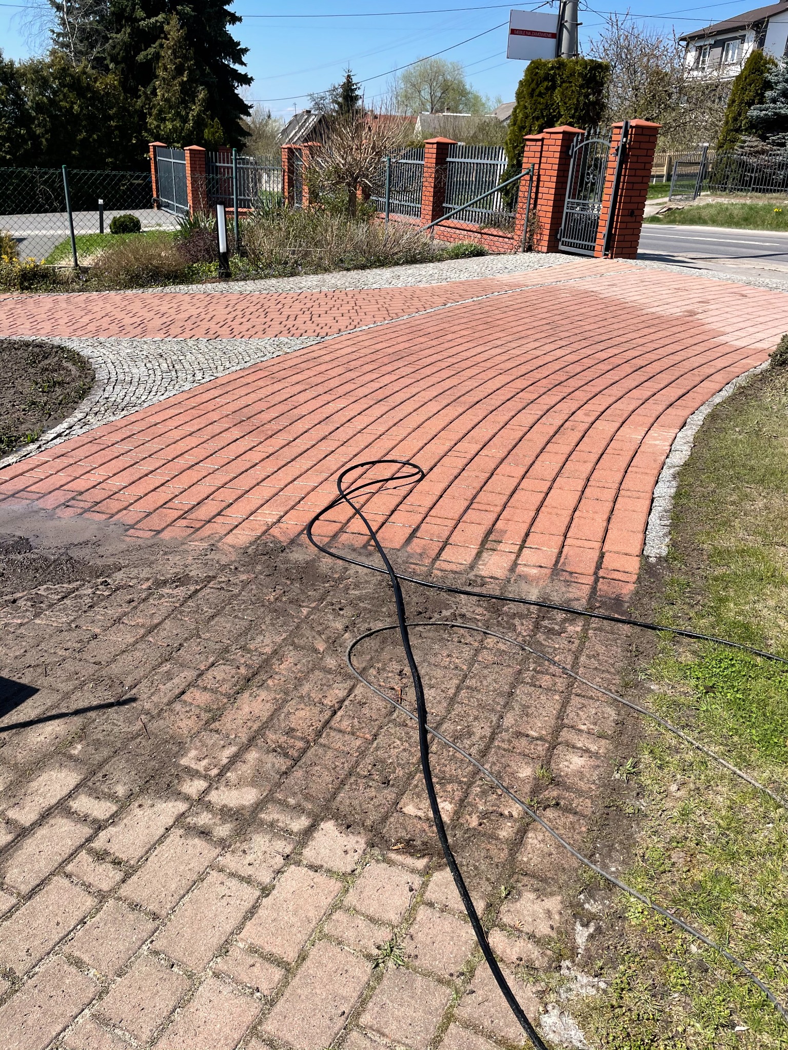 Częściowo oczyszczona, zakrzywiona ścieżka z czerwonej kostki brukowej z widocznymi śladami brudu i wężem ciśnieniowym, prowadząca do bramy z ceglanymi słupkami w słoneczny dzień.