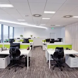 Nowoczesne biuro z białymi biurkami, czarnymi krzesłami obrotowymi i zielonymi przegrodami, oświetlone światłem dziennym i sztucznym, z kolumną po lewej stronie i wykładziną dywanową w odcieniach...