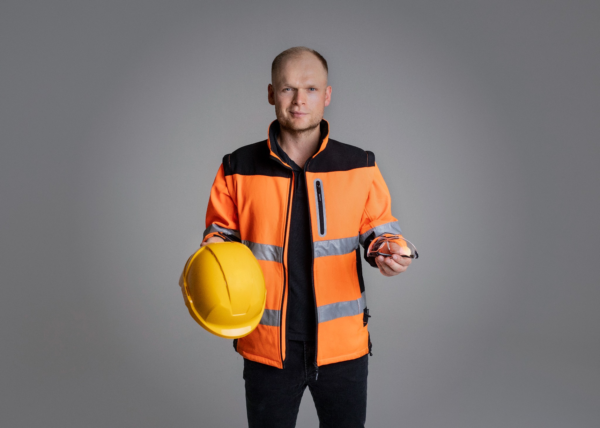 Mężczyzna w odblaskowej kamizelce trzyma żółty kask i okulary ochronne na szarym tle. Kadr centralny.