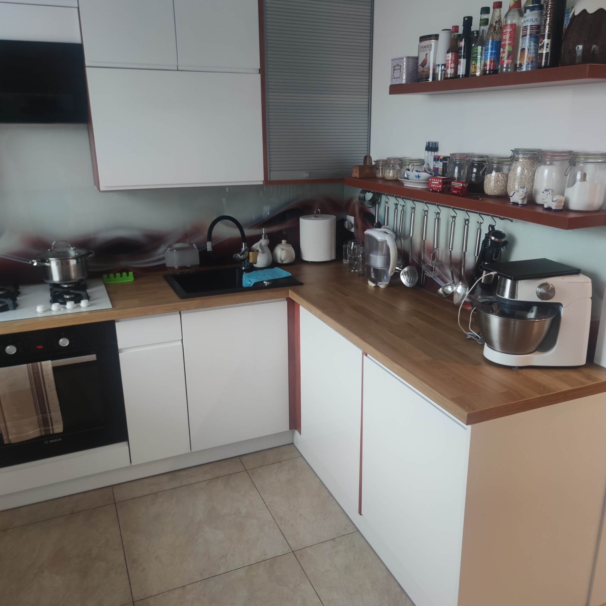 Nowoczesna kuchnia z białymi szafkami, drewnianym blatem, czarnym zlewem i indukcyjną płytą grzewczą z garnkiem, widok na półki z przyprawami i robot kuchenny.