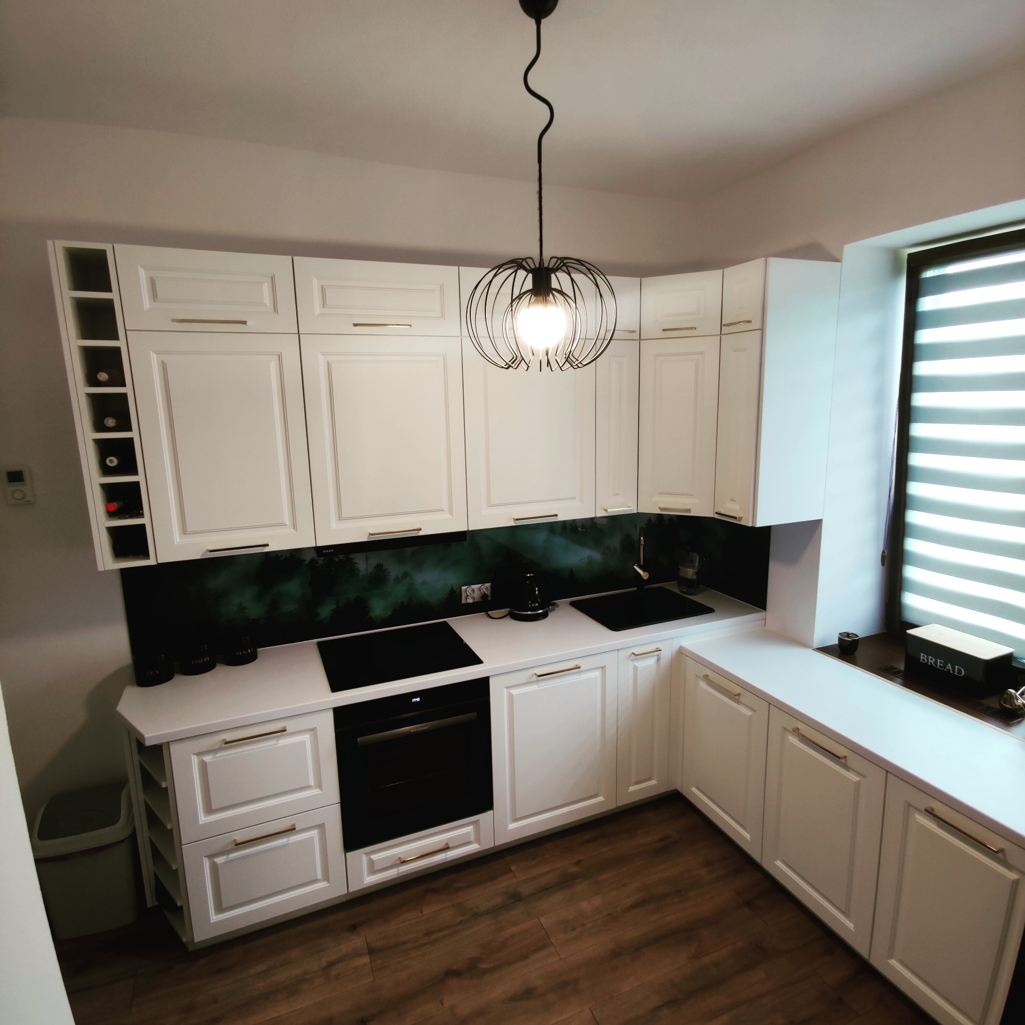 Nowoczesna kuchnia w bieli z drewnianą podłogą, czarnym blatem i szklanym panelem ściennym z motywem leśnym, oświetlona designerską lampą wiszącą.