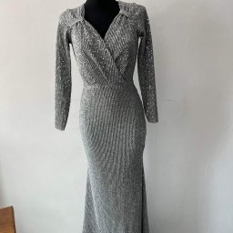 by K&K-sklep odzieżowy - Długa, srebrna suknia wieczorowa z długim rękawem, drapowanym dekoltem w kształcie litery V, prezentowana na czarnym manekinie na tle białej ściany i szarej podłogi.