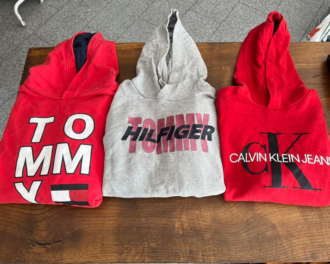 Trzy złożone bluzy z kapturem: czerwona Tommy Hilfiger, szara Tommy Hilfiger Downhill Challenge i czerwona Calvin Klein Jeans, ułożone na drewnianym stole.