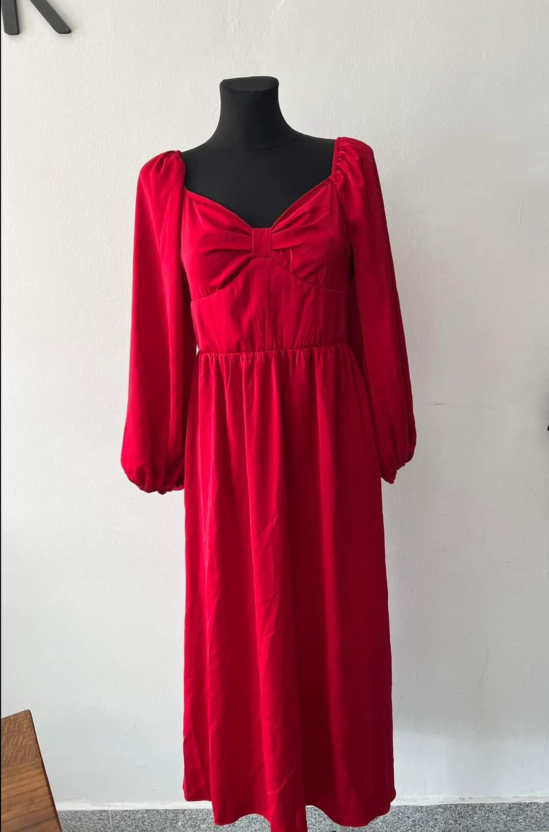 Czerwona, welurowa sukienka midi z długimi, bufetowymi rękawami i dekoltem w kształcie serca, prezentowana na czarnym manekinie na tle białej ściany.