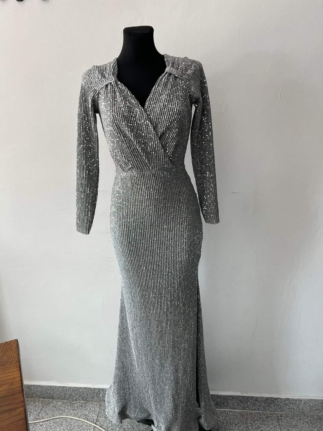 Długa, srebrna suknia wieczorowa z długim rękawem, drapowanym dekoltem w kształcie litery V, prezentowana na czarnym manekinie na tle białej ściany i szarej podłogi.