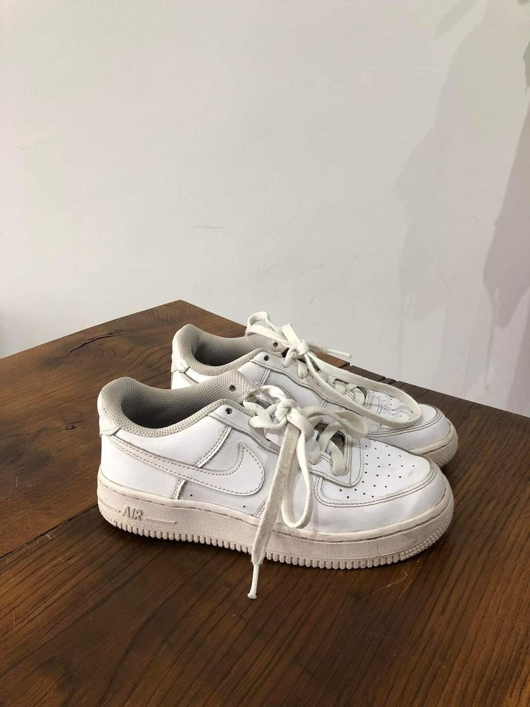 Para białych, lekko zabrudzonych butów sportowych Nike Air Force 1 na drewnianym stole, sznurówki rozwiązane.