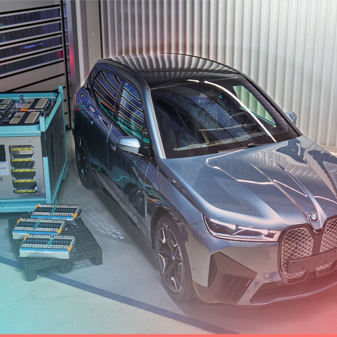 Szary elektryczny SUV marki BMW z widocznymi akumulatorami na paletach i stacją ładowania w tle, w jasnym garażu.