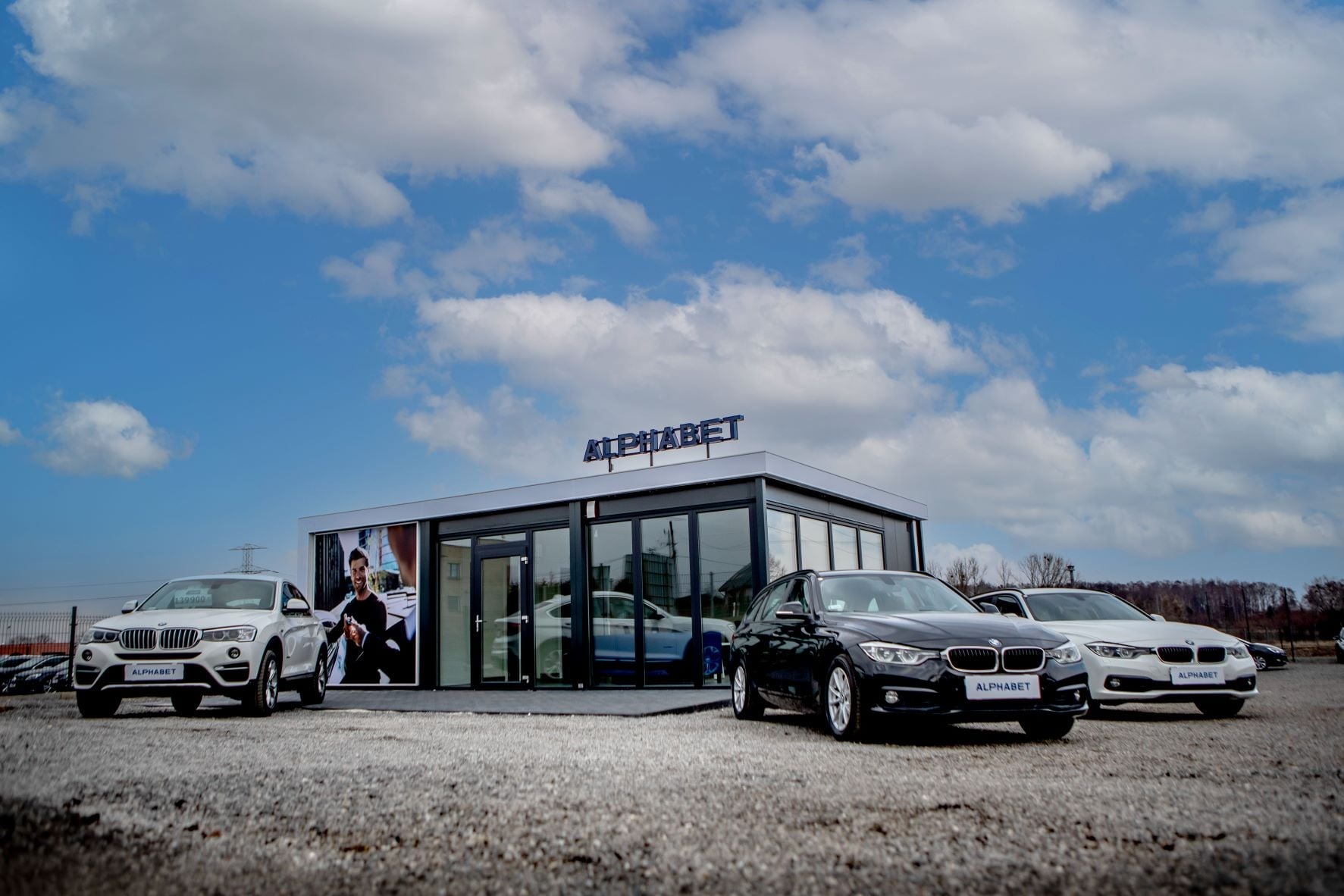 Nowoczesny pawilon z logo ALPHABET, przed którym stoją zaparkowane samochody BMW w kolorach białym i czarnym, na żwirowym placu pod błękitnym niebem z chmurami.