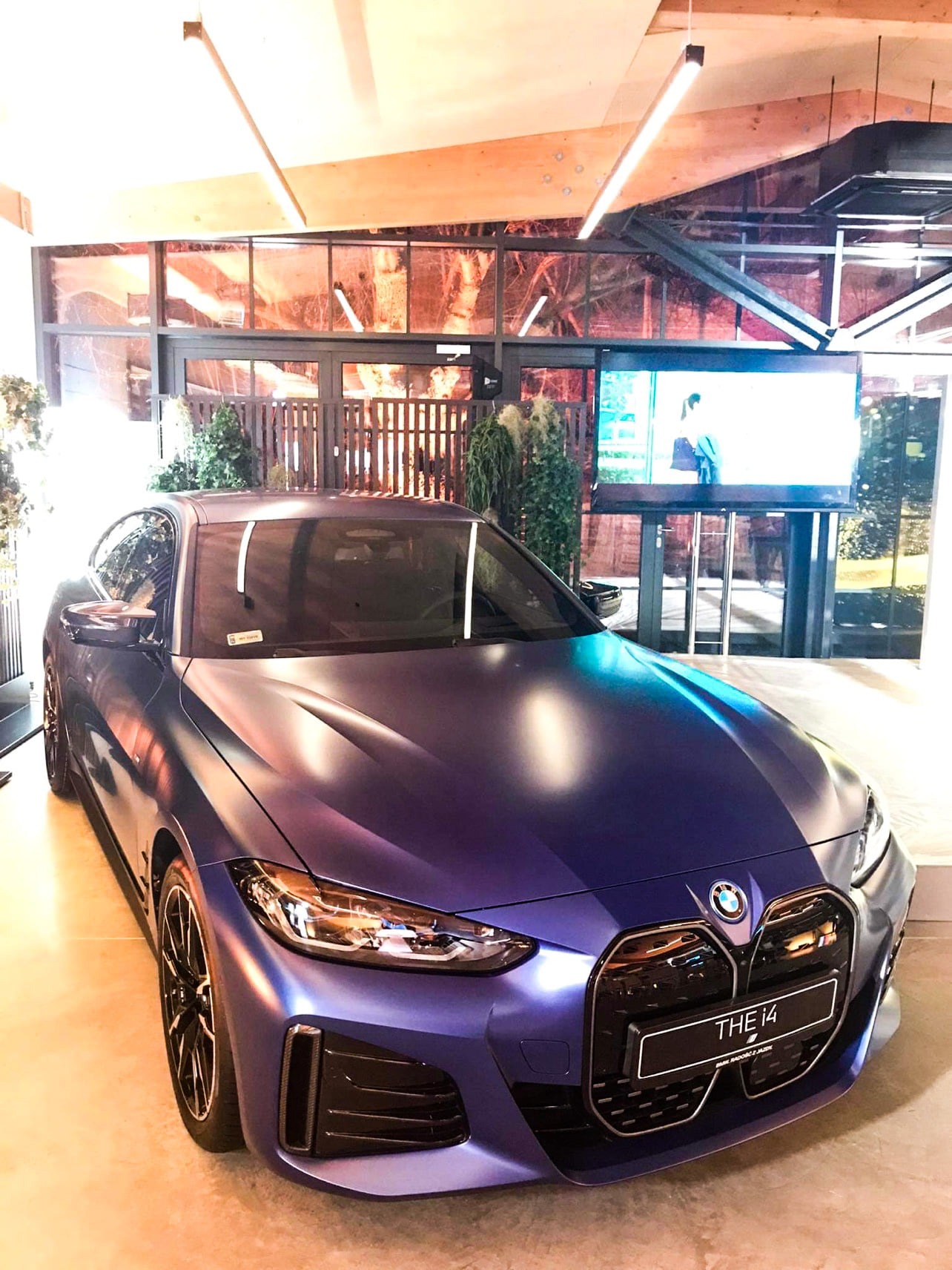 Matowy, fioletowy samochód BMW i4 prezentowany w salonie z drewnianym sufitem i ekranem wyświetlającym w tle.