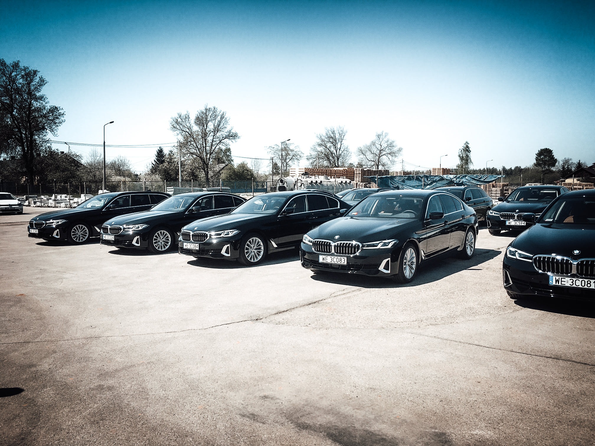 Rząd czarnych samochodów BMW serii 5 zaparkowanych na betonowym placu w słoneczny dzień, z widocznymi numerami rejestracyjnymi i drzewami w tle.
