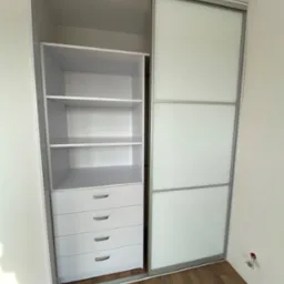 Wnętrze szafy wnękowej z przesuwnymi, szklanymi drzwiami, widoczne p&oacute;łki i szuflady, na podłodze panele drewnopodobne, obok ściana z gniazdkiem elektrycznym.