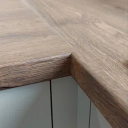 Fragment narożnika blatu z ciemnego drewna o widocznej strukturze słoj&oacute;w, zamontowanego na szafkach kuchennych w odcieniach szarości.