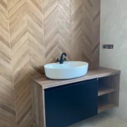 Nowoczesna łazienka z umywalką nablatową, czarną armaturą i szafką z drewnianym blatem oraz czarnym frontem, tło stanowi ściana wyłożona drewnopodobną płytką w jodełkę.