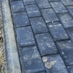 Ułożona, prostokątna, szara kostka brukowa z widocznymi śladami piasku i ziemi na powierzchni, ułożona ukośnie do kadru, z widocznym obrzeżem.