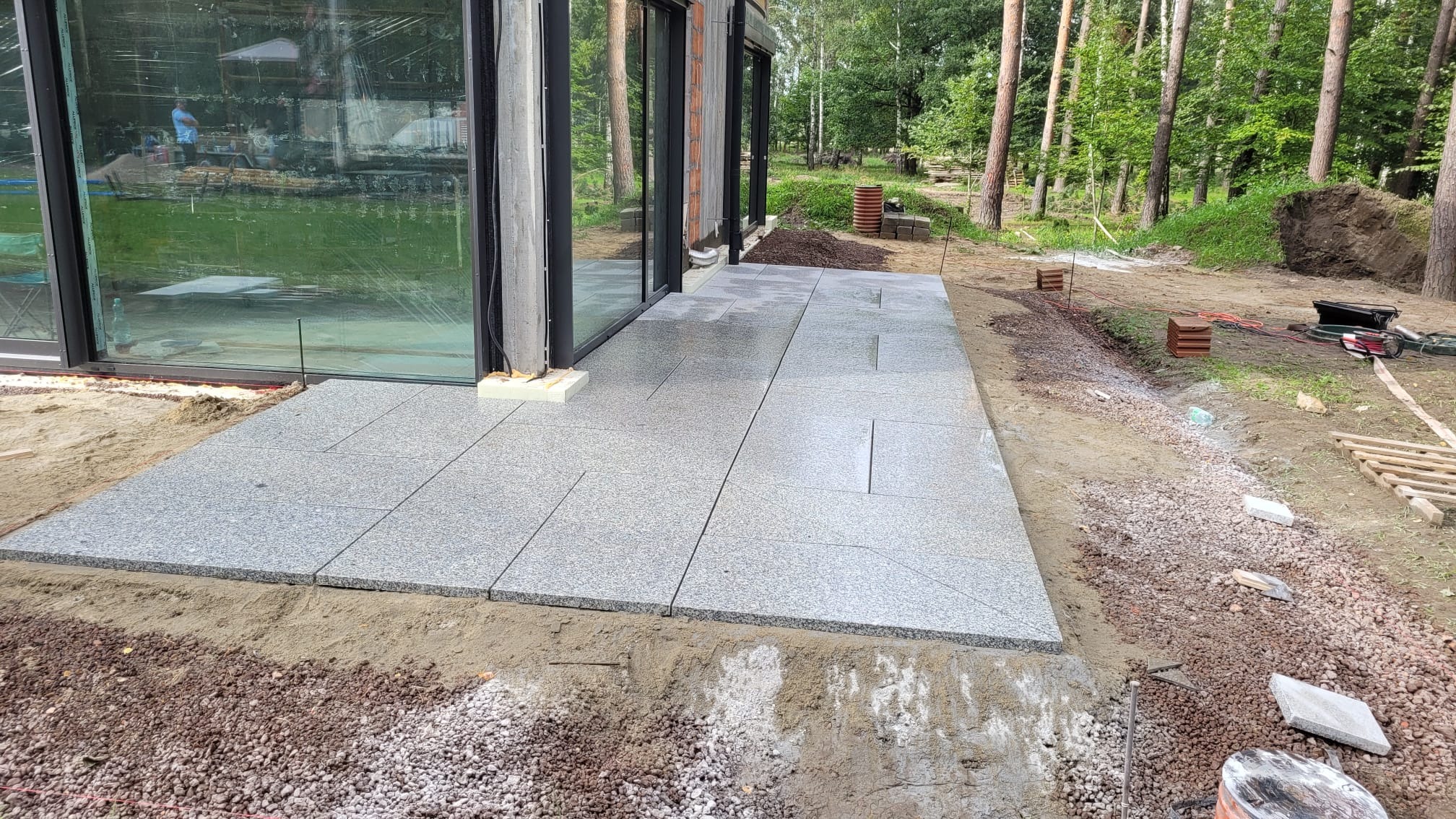 Świeżo ułożony, szary taras z płyt granitowych przylegający do budynku z dużymi oknami, otoczony przygotowanym podłożem z piasku i żwiru, z widocznymi narzędziami i materiałami budowlanymi w tle.