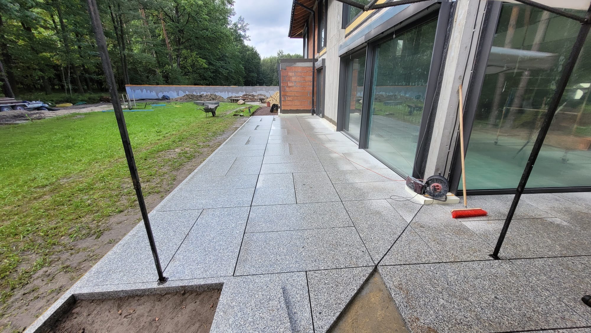 Nowo ułożony taras z jasnoszarej kostki granitowej przy nowoczesnym domu z dużymi oknami, widoczne narzędzia pracy i przygotowane podłoże pod schody.