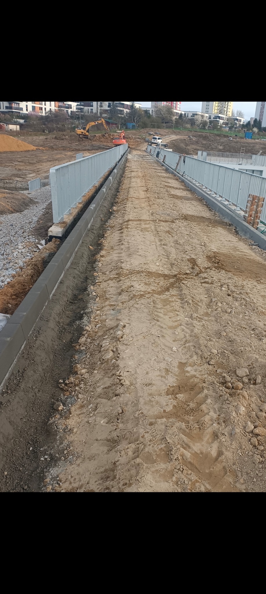 Budowa drogi: widok na przygotowany grunt pod nawierzchnię, z widocznymi obrzeżami betonowymi i metalowymi barierkami. W tle sprzęt budowlany i zabudowa miejska.