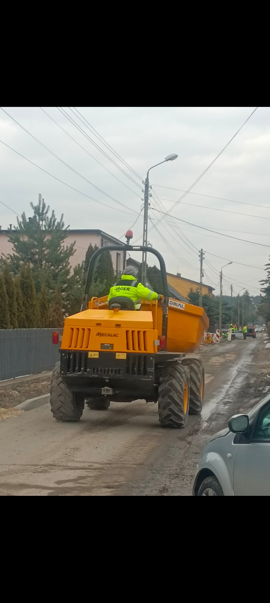 Żółta wywrotka Mecalac na błotnistej drodze podczas budowy, w tle widoczni pracownicy w odblaskowych kamizelkach i znaki drogowe.