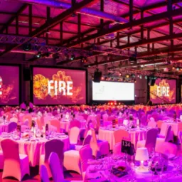 Sala bankietowa z okrągłymi stołami i krzesłami w pokrowcach, oświetlona na różowo, z dużymi ekranami wyświetlającymi napis 'FIRE' z efektem płomieni.