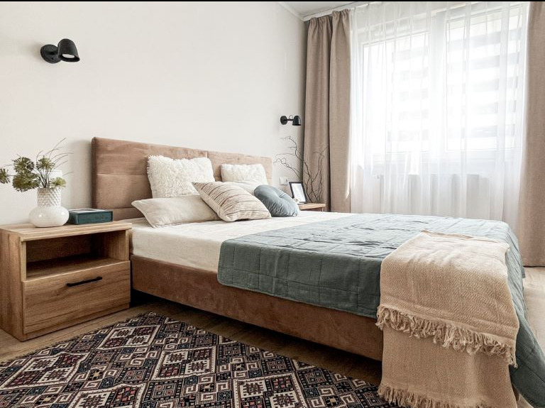 Przytulna sypialnia po home stagingu z beżowym łóżkiem, drewnianą szafką nocną z wazonem i wzorzystym dywanem, oświetlona światłem dziennym przez okno z jasnymi zasłonami.