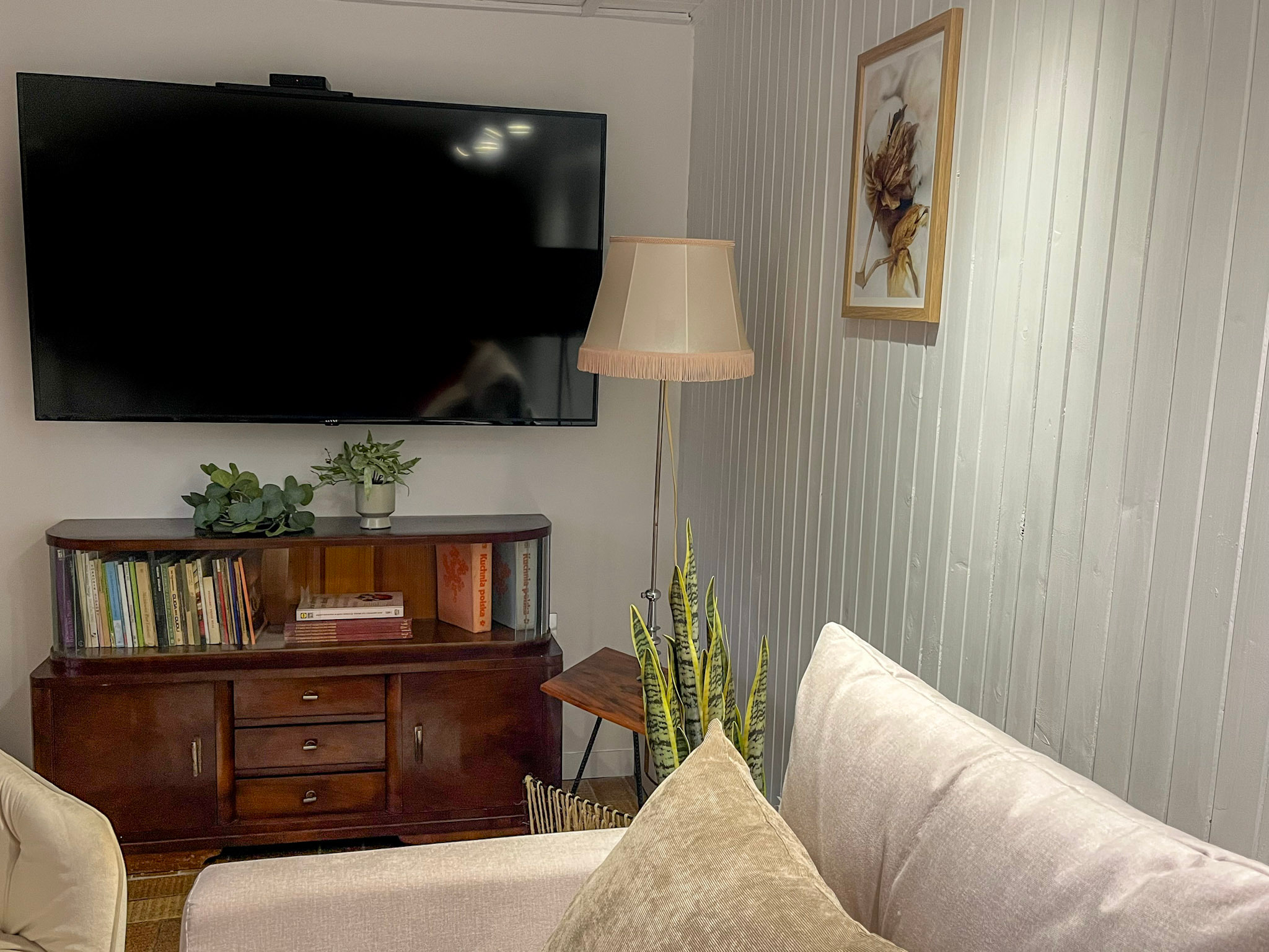 Salon po metamorfozie: brązowy kredens z książkami pod telewizorem, lampa podłogowa z beżowym abażurem, sofa z poduszkami i obraz na ścianie z boazerii.