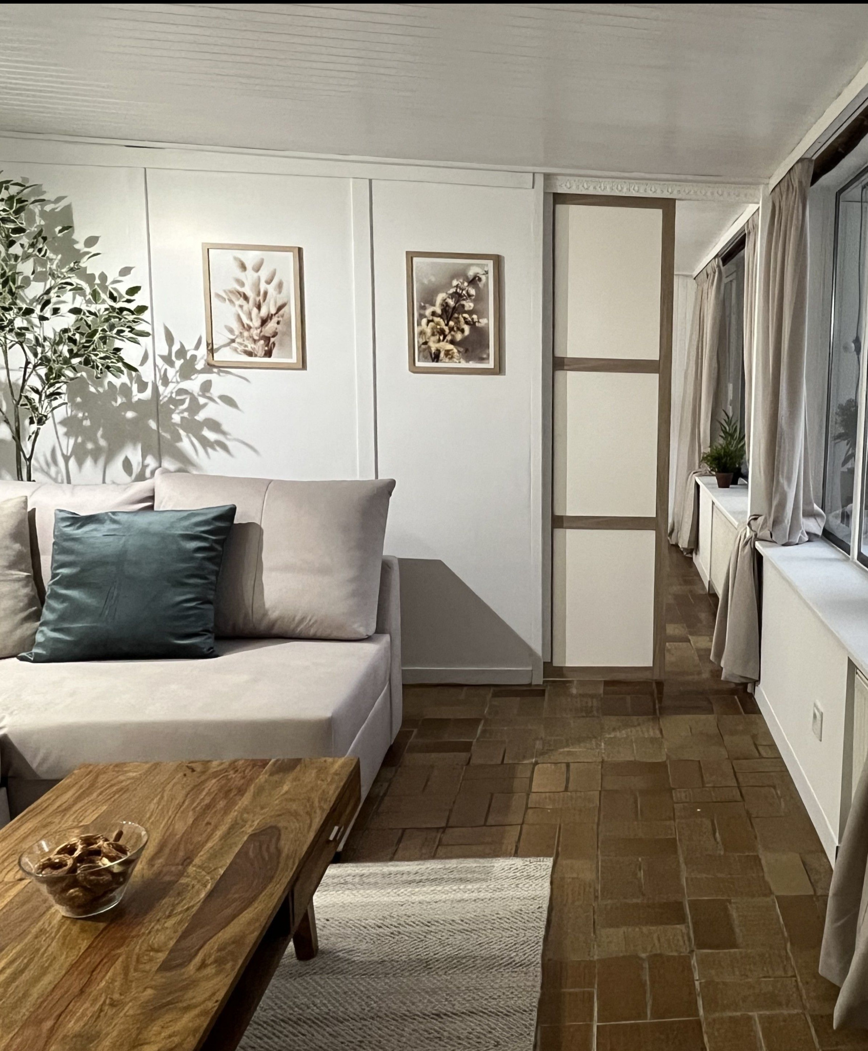 Jasny salon po metamorfozie: beżowa sofa z zieloną poduszką, drewniany stolik kawowy z przekąskami, obrazy na ścianie i oryginalna podłoga z cegiełek.