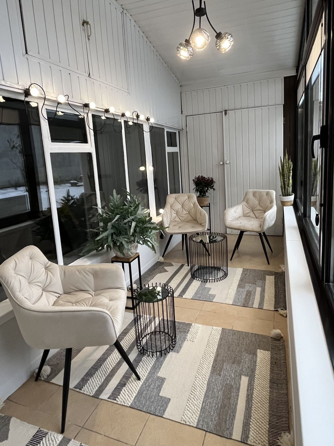 Jasny hol po metamorfozie: kremowe fotele, geometryczny dywan, rośliny w donicach, dekoracyjne oświetlenie. Widok na zewnątrz przez okna.