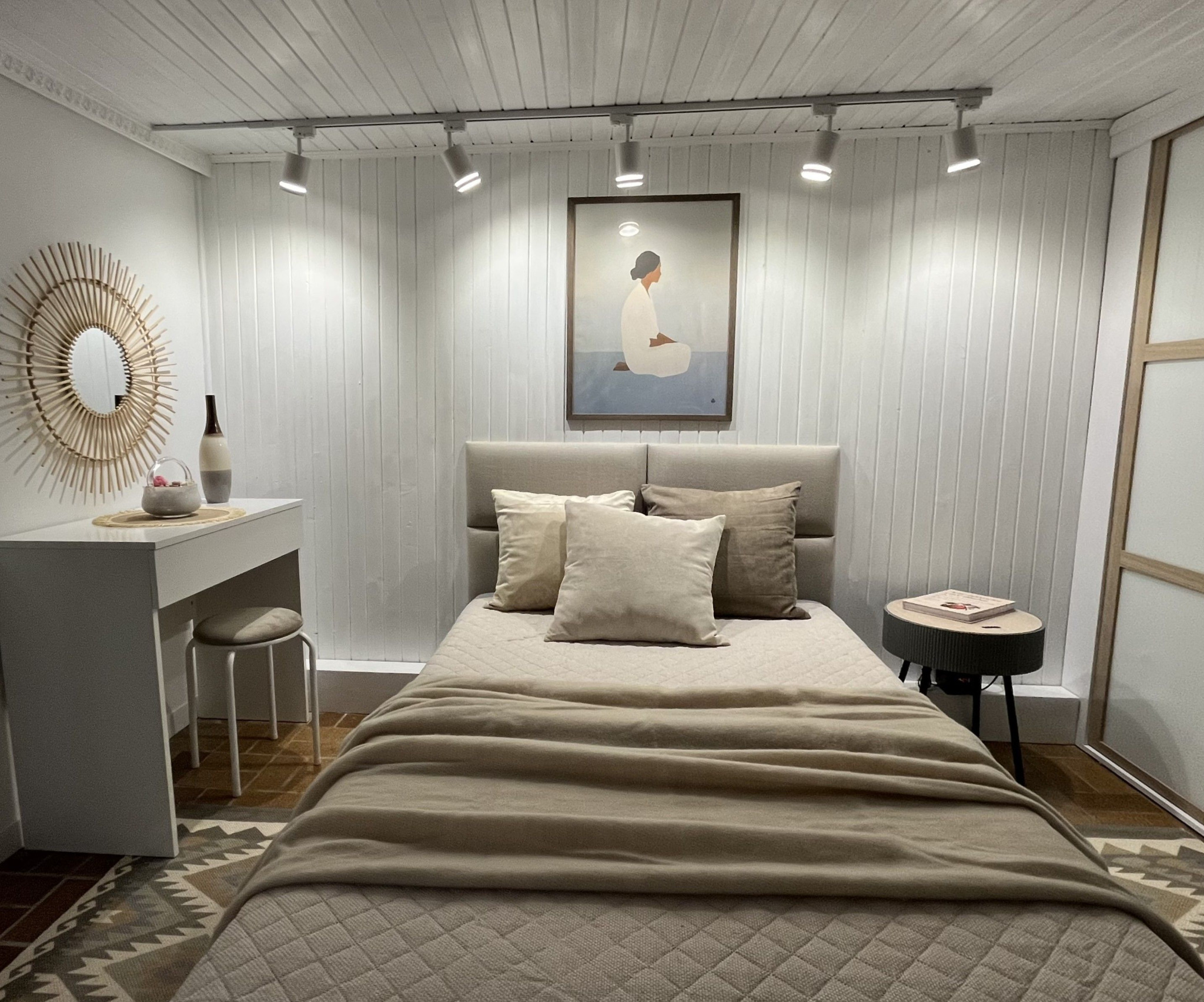 Jasna sypialnia po metamorfozie: beżowe tekstylia, drewniane panele ścienne, okrągłe lustro z rattanu i minimalistyczny obraz nad łóżkiem. Przytulne oświetlenie punktowe.