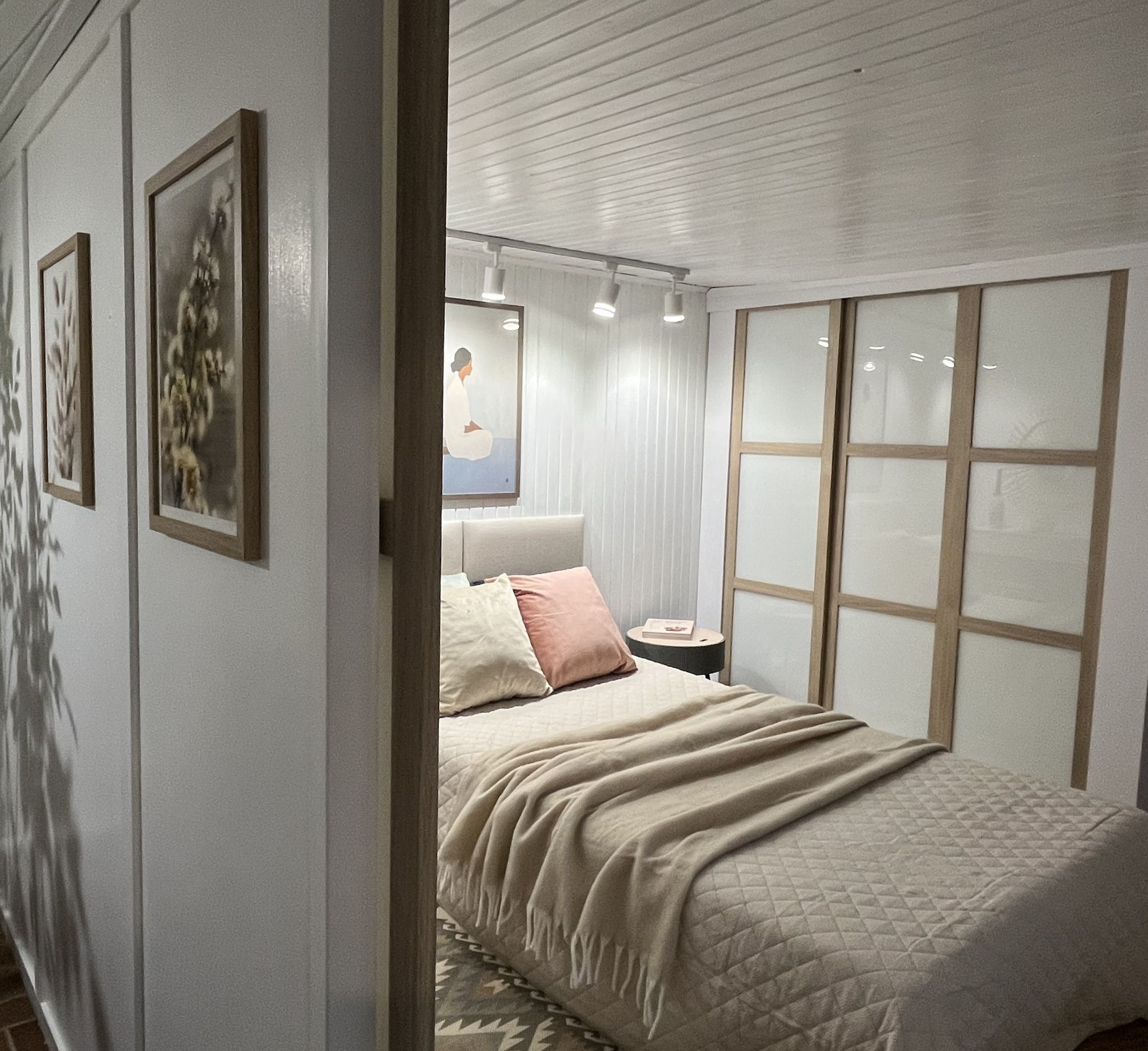 Jasna sypialnia po metamorfozie: beżowa narzuta na łóżku, obrazy na ścianie i przesuwne drzwi szafy w tle. Widok zza framugi drzwi.