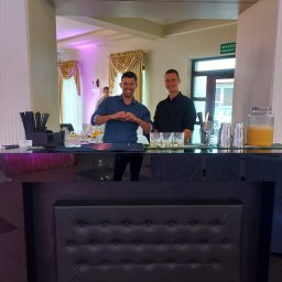 Stanisław Kalinowski Bohema - Uśmiechnięci barmani za czarnym, eleganckim barem z pikowanym frontem, przygotowani do obsługi gości na tle sali bankietowej z dekoracyjnymi zasłonami.