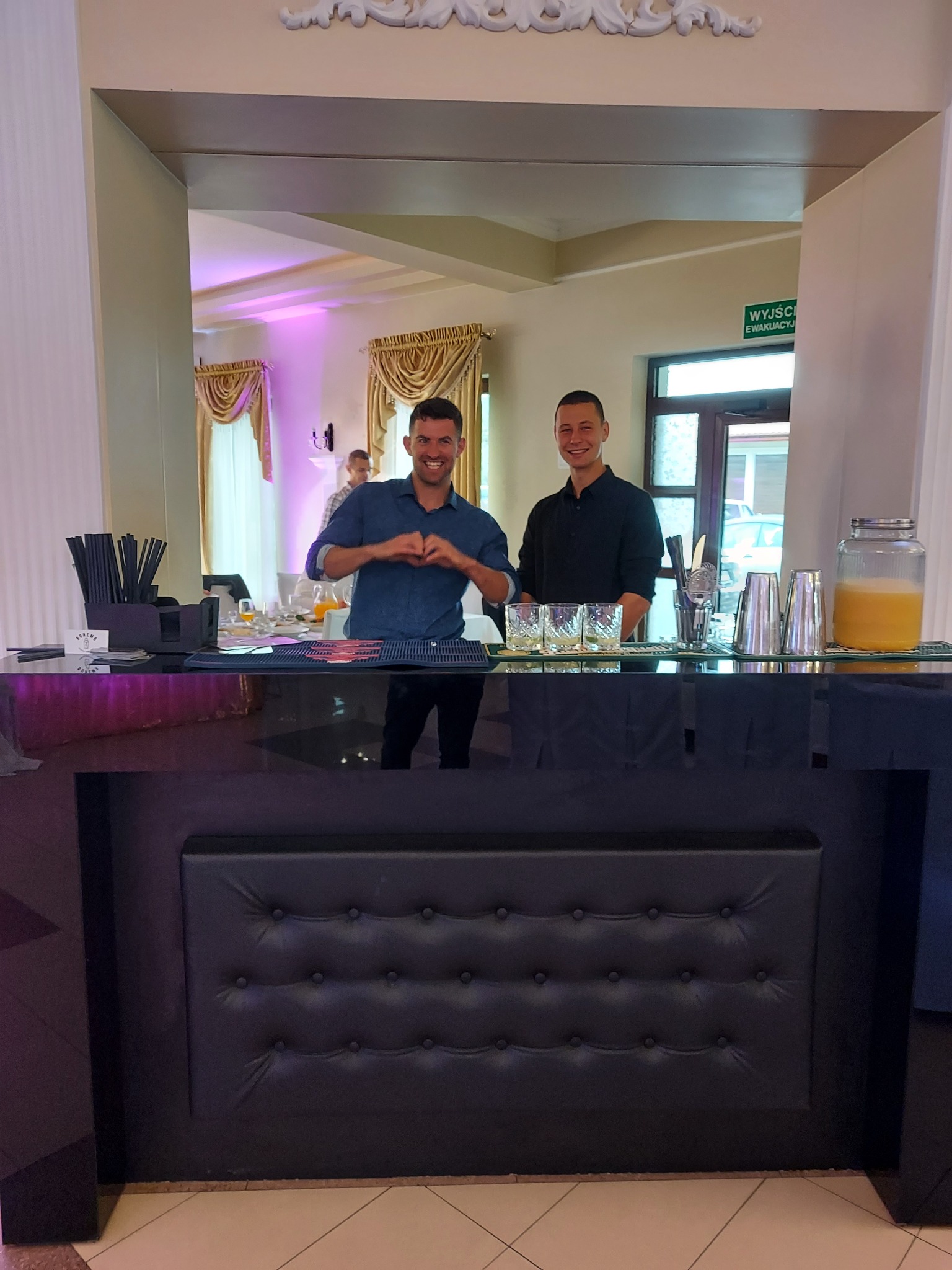 Uśmiechnięci barmani za czarnym, eleganckim barem z pikowanym frontem, przygotowani do obsługi gości na tle sali bankietowej z dekoracyjnymi zasłonami.