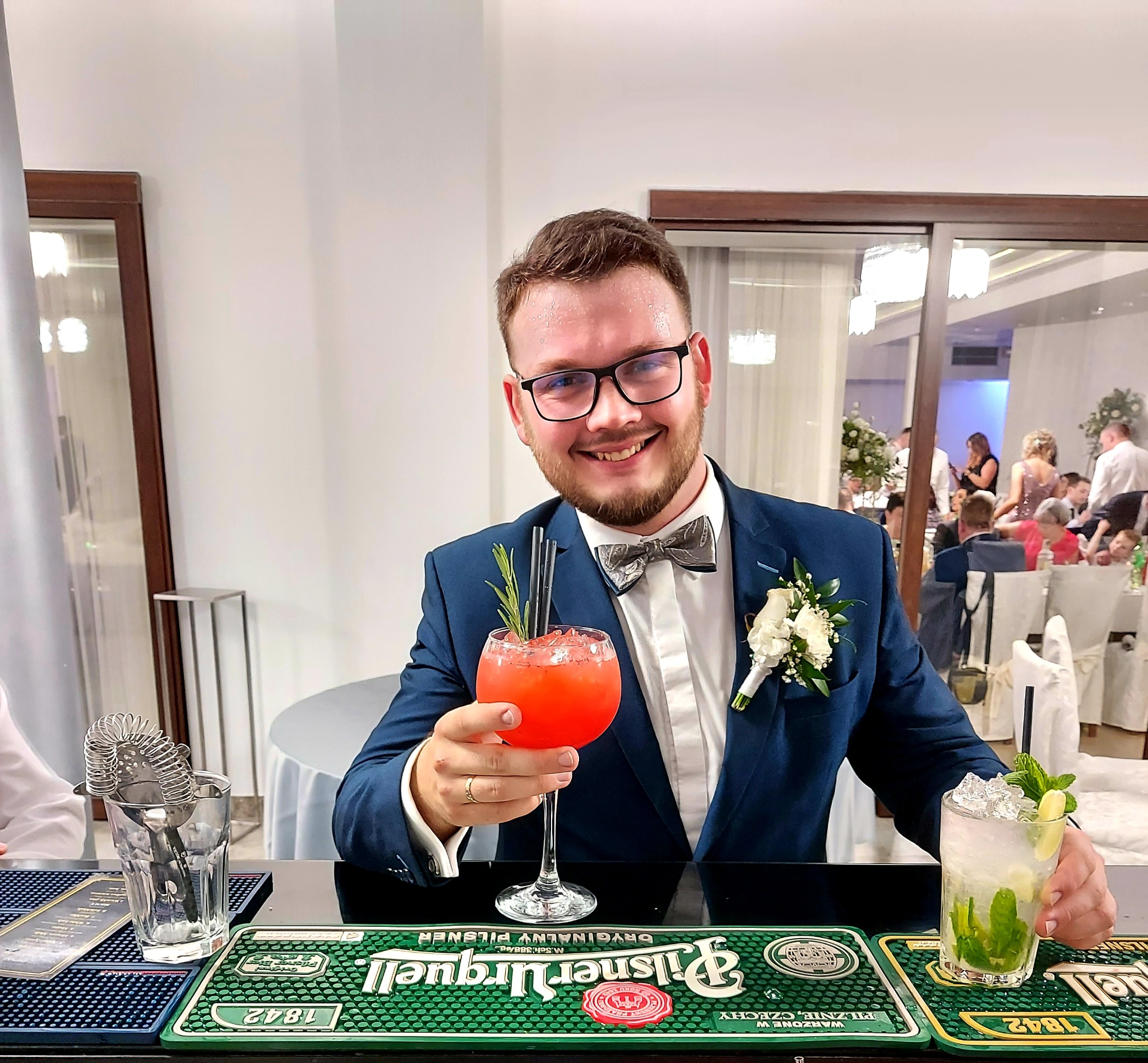 Uśmiechnięty barman w eleganckim stroju, prezentuje dwa kolorowe koktajle na tle sali bankietowej podczas przyjęcia.